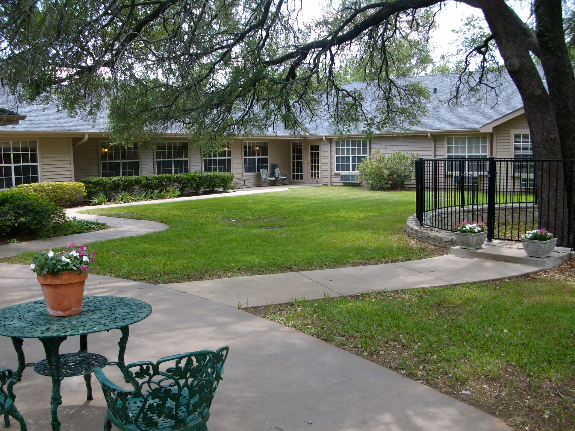 Outdoor courtyard area of a senior living facility with a green metal table and chairs, a potted plant on the table, a curved concrete walkway, green grass, trees providing shade, and a beige building with multiple windows in the background.
