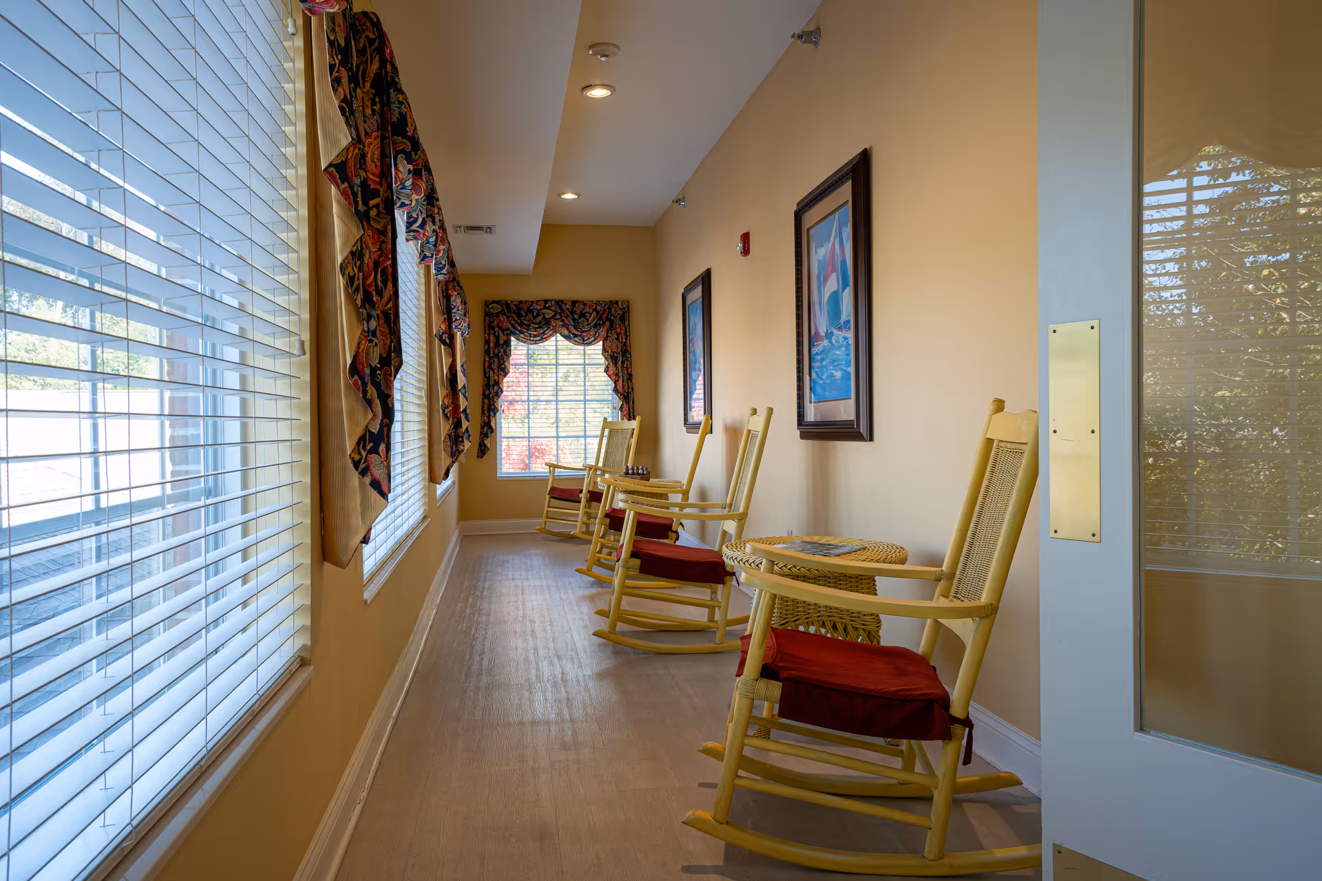 A bright hallway with large windows on the left side covered with blinds and decorative valances. On the right side, there are several yellow wooden rocking chairs with red cushions and a small wicker table. The walls are painted light yellow and decorated with framed nautical-themed artwork. The floor is light-colored wood, and recessed ceiling lights illuminate the space.