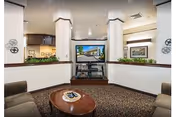 Interior view of a common area in a retirement community featuring two beige armchairs facing a flat-screen TV mounted on a stand between two white pillars. The room has patterned carpet flooring, a wooden coffee table with a decorative plate, and framed artwork on the walls. There are green plants on ledges behind the pillars.