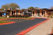 Exterior view of a single-story nursing home building with large windows and brick walls. The entrance is visible with a covered walkway, surrounded by shrubs and a parking area with marked spaces and red curbs.