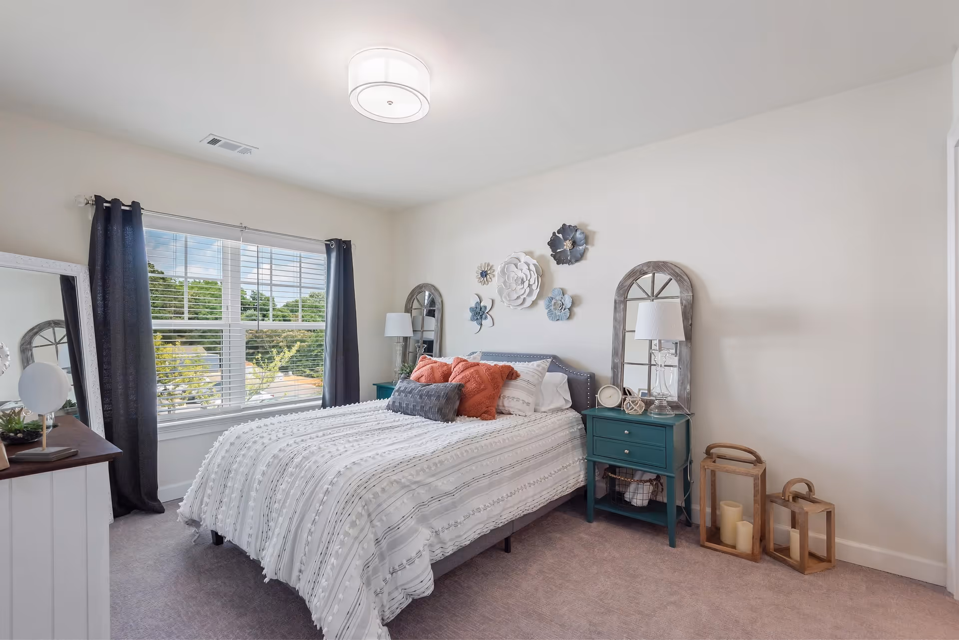 A bright bedroom with a large window covered by dark curtains, a bed with white textured bedding and multiple pillows in white, gray, and coral colors. There are two teal nightstands on either side of the bed, each with a lamp and decorative items. The wall above the bed is adorned with large flower-shaped wall art. Two wooden lanterns with candles are placed on the floor next to one nightstand. A full-length mirror leans against the wall near the window.