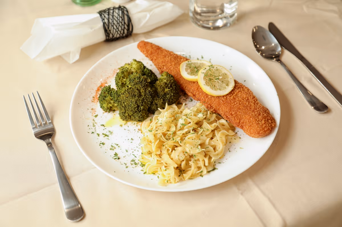A plate of food on a beige tablecloth featuring breaded fish fillet garnished with two lemon slices, steamed broccoli, and buttered noodles sprinkled with herbs. A fork is placed to the left of the plate, and a spoon and knife are to the right. A folded white napkin with a black napkin ring and a glass of water are also visible on the table.