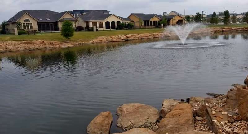 A pond with a water fountain in the center, surrounded by rocks and grass. In the background, there are several single-story buildings with beige walls and dark roofs under a cloudy sky.