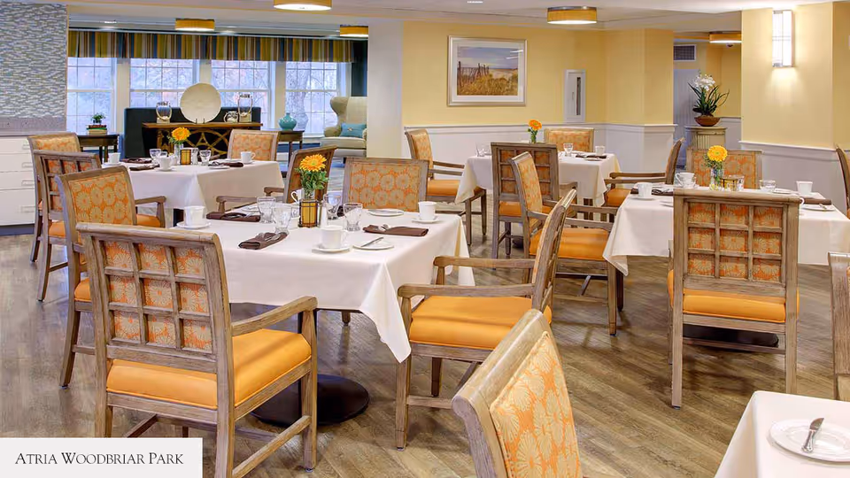 A bright dining room with several tables covered in white tablecloths, each set with cups, glasses, plates, and silverware. The chairs have wooden frames with orange cushioned seats and patterned backs. There are yellow flowers in small vases on each table. The room has large windows with striped valances, light yellow walls, and wooden flooring. A framed picture hangs on the wall, and there are decorative plants in the background.
