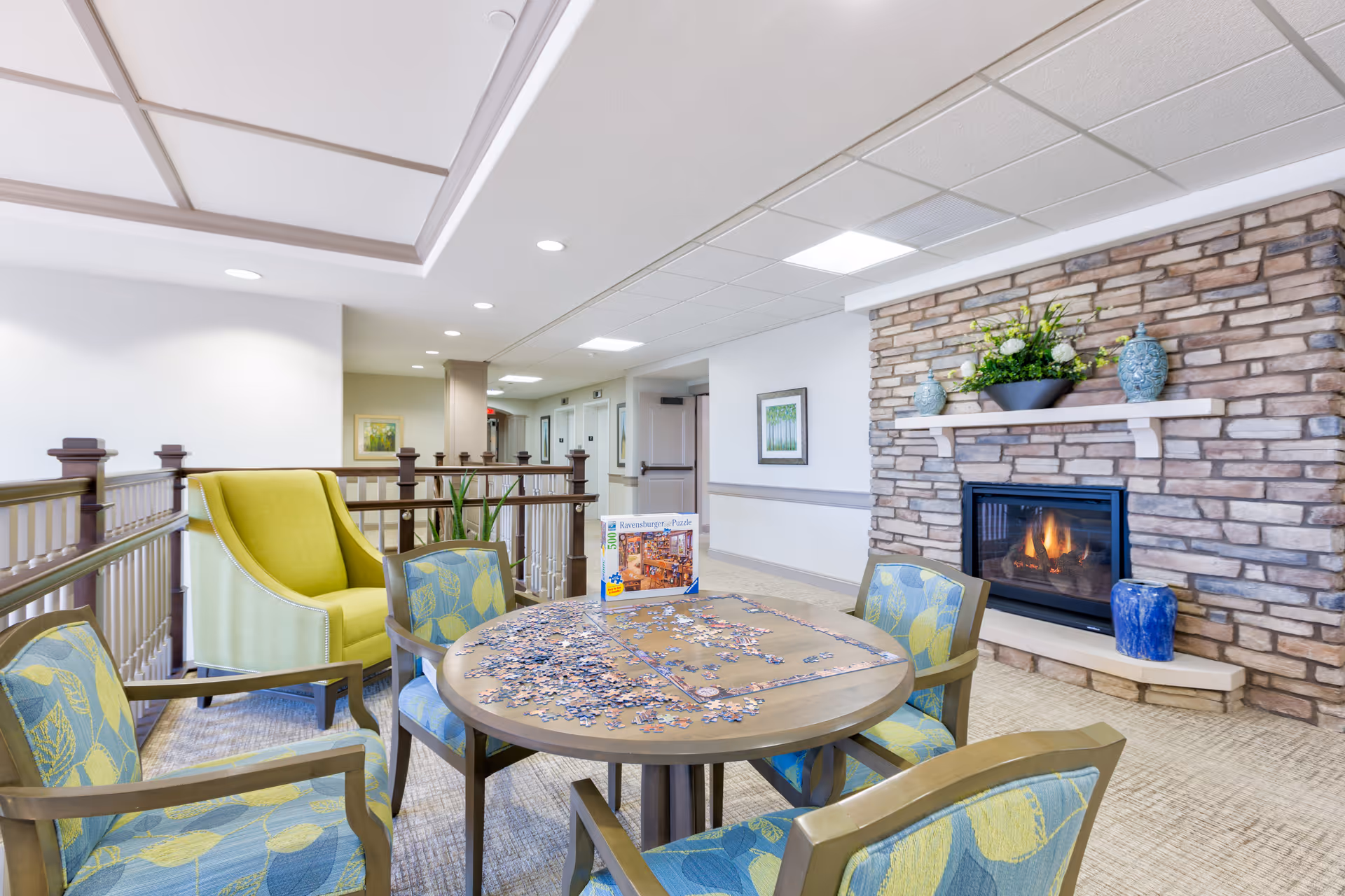 A cozy senior living common area with a round table in the center, partially completed puzzle pieces spread on the table, surrounded by four chairs with blue and green leaf-patterned upholstery. A green armchair is positioned near a wooden railing. A stone fireplace with a lit fire is on the right wall, decorated with vases and a flower arrangement on the mantel. The space is well-lit with recessed ceiling lights and has framed artwork on the walls.