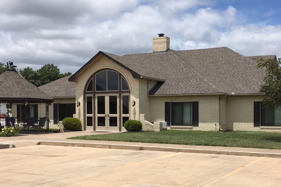 Exterior view of a single-story brick building with a large arched window and double glass doors at the entrance. There is a small gazebo with seating on the left side and a parking lot in the foreground. The sky is partly cloudy.