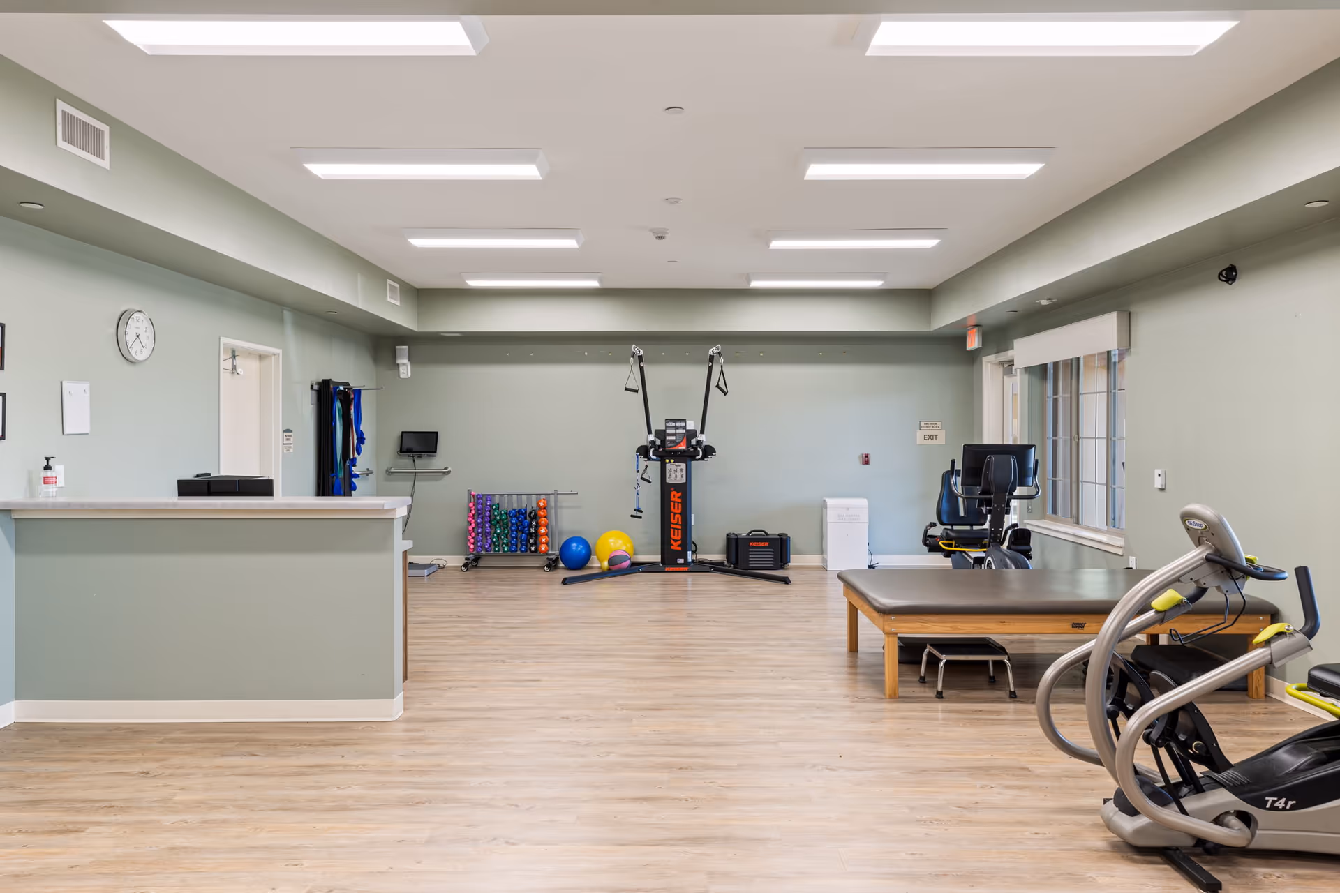 A spacious exercise room with light green walls and wood flooring. The room contains various fitness equipment including a Keiser exercise machine, colorful exercise balls, a rack of small weights, a recumbent bike, and a padded exercise table. There is a reception counter on the left side, a clock on the wall, and a window on the right side letting in natural light.
