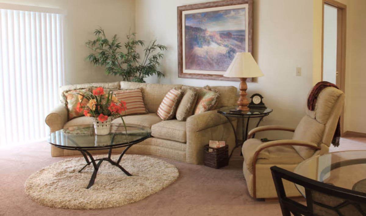 Well-lit living room with a beige sofa and recliner, glass coffee table on a round rug, side table with lamp, framed artwork, and a potted plant.