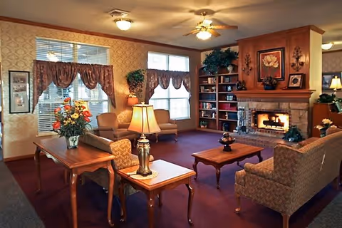 A cozy living room with a lit fireplace, wooden bookshelves filled with books and decorative items, two large windows with valance curtains, multiple upholstered chairs and sofas, a wooden coffee table, and a table lamp on a side table. The walls have patterned wallpaper and there is a ceiling fan with a light fixture.