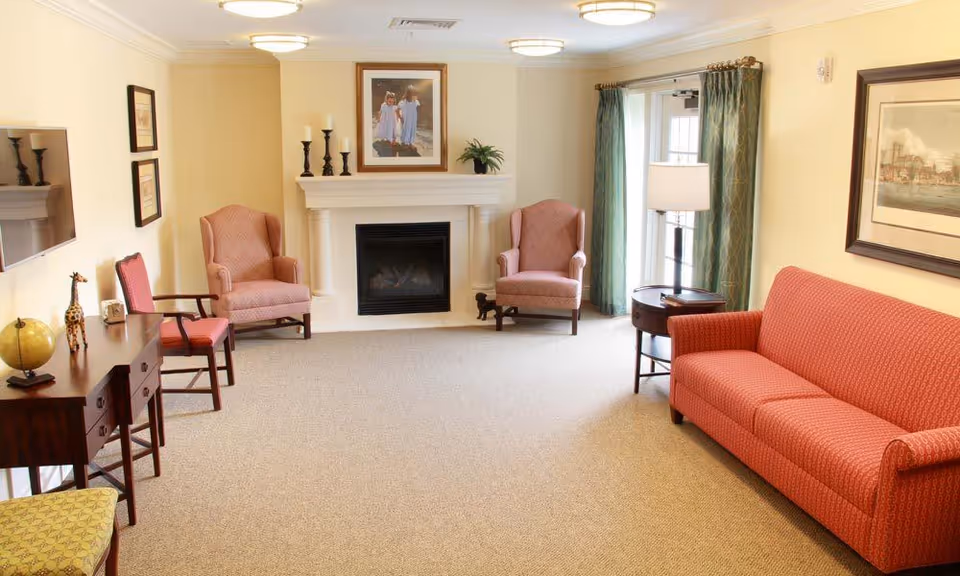 A cozy living room with beige walls and carpet, featuring a red patterned sofa on the right, two matching armchairs near a white fireplace with candles and a framed picture above it, a small round side table with a lamp, a wooden desk with a globe and giraffe figurine, and a window with green curtains letting in natural light.