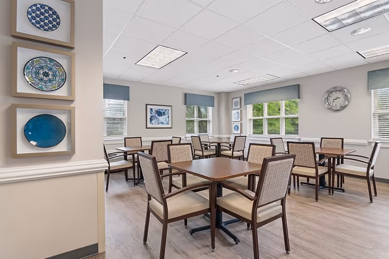 A bright dining room with several wooden tables and cushioned chairs arranged neatly. The room has large windows with blue valances allowing natural light to enter. Decorative plates and framed artwork adorn the walls, and a clock is mounted on one wall.