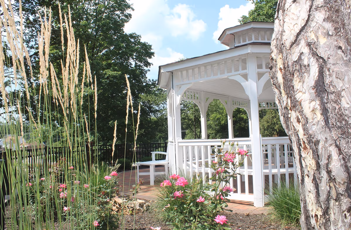 A white wooden gazebo surrounded by green trees, tall ornamental grasses, and pink flowering plants under a blue sky with some clouds. A large tree trunk is visible on the right side of the image.