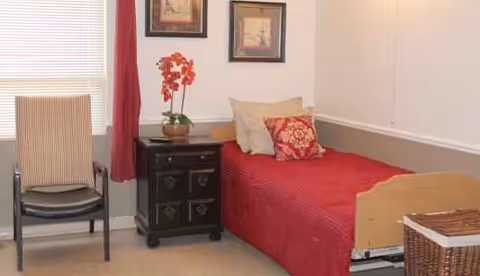 A cozy bedroom with red bedspread, nightstand, chair, and window blinds in a senior facility.