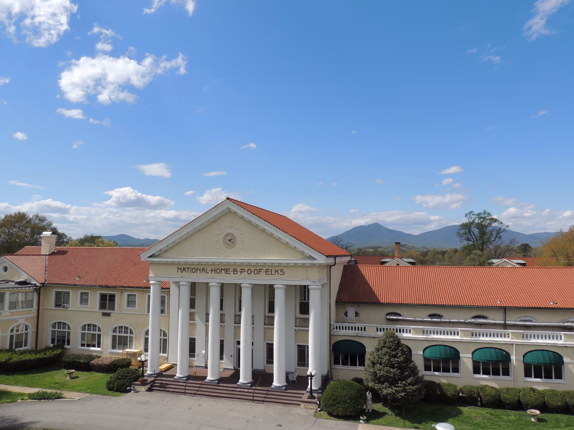 Front exterior of a large neoclassical building with tall white columns, a red tile roof, and mountains in the background.