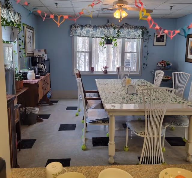 A dining room with a rectangular table covered with a floral tablecloth surrounded by white chairs with tennis balls on the legs. The room has light blue walls, a window with a floral valance, and a ceiling fan with lights. There are colorful paper chains hanging from the ceiling. A wooden sideboard with a coffee maker and plants is on the left side of the room.