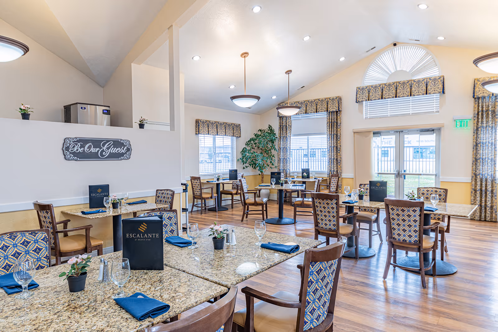 Bright senior living dining room with multiple set tables, patterned chairs, and large windows.