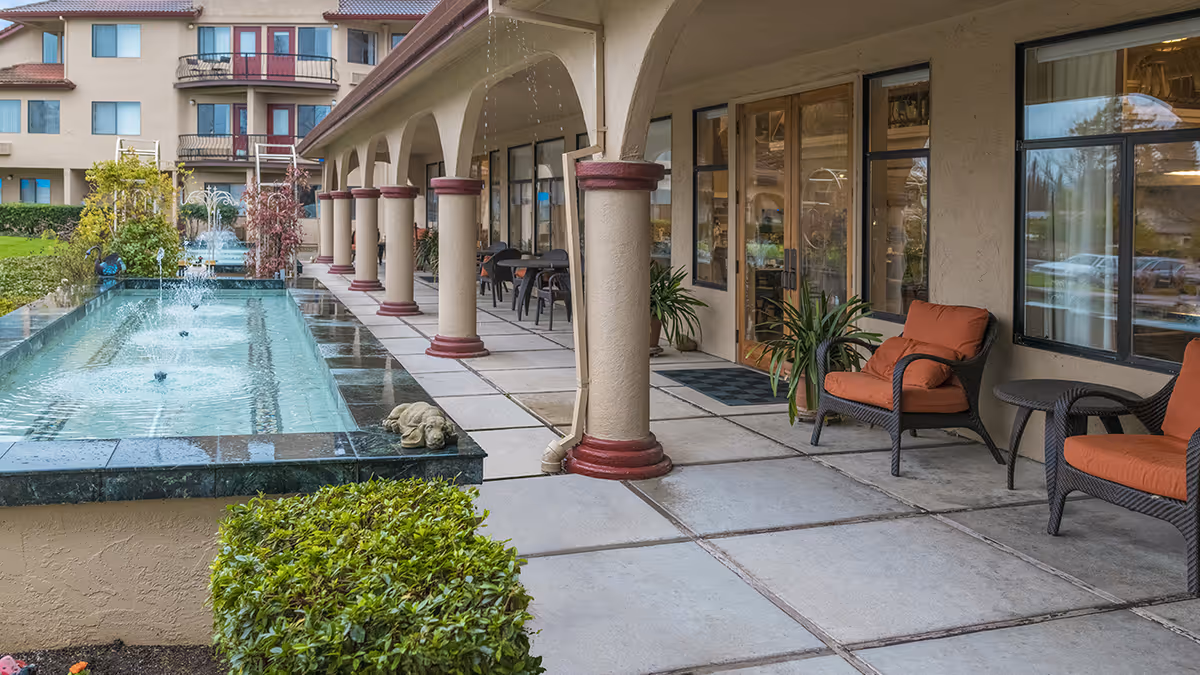Outdoor covered walkway with arched columns, a long fountain, and cushioned patio seating outside the building.