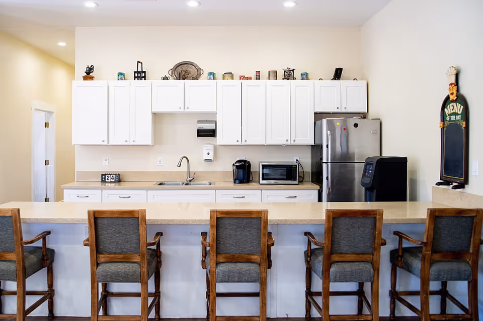 A clean and modern kitchen area with white cabinets, a stainless steel refrigerator, a microwave, a coffee maker, and a sink. There is a long counter with five wooden chairs with gray cushions. On the wall to the right, there is a decorative menu board shaped like a chef.