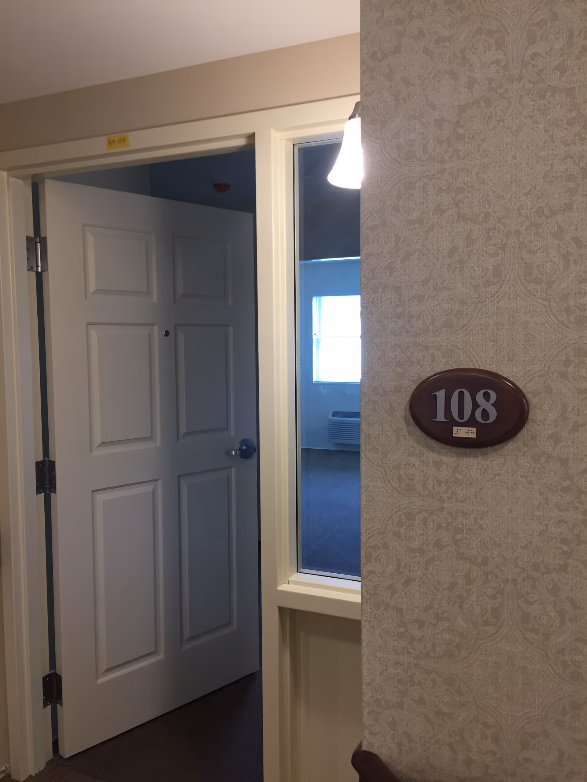 An open apartment door in a hallway with a patterned wall and a room number plaque reading 108.