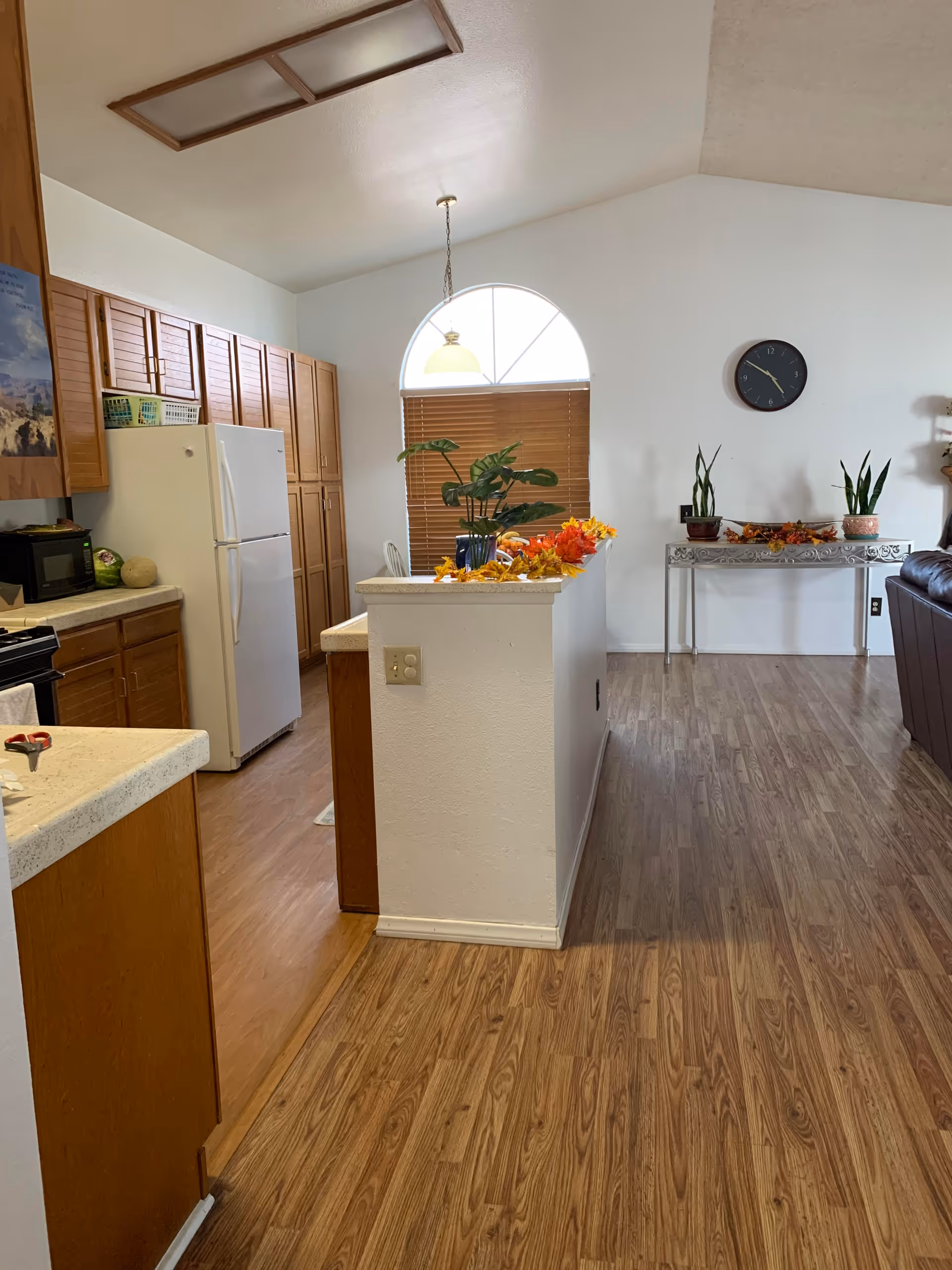 Interior view of a kitchen and living area with wooden cabinets, a white refrigerator, a microwave, and a countertop decorated with autumn leaves and plants. There is a window with a half-circle top and blinds, a hanging light fixture, a wall clock, and a small table with potted plants against a white wall. The floor is wood laminate.
