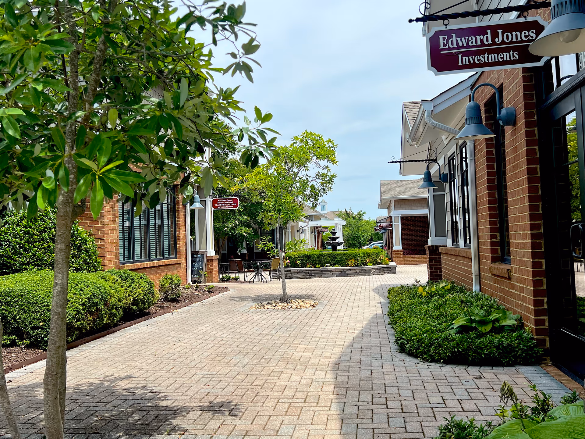 A paved outdoor walkway lined with small trees and bushes on both sides, flanked by brick buildings with signs for businesses including Edward Jones Investments. There is a small seating area with a table and chairs, and a fountain is visible in the background under a partly cloudy sky.