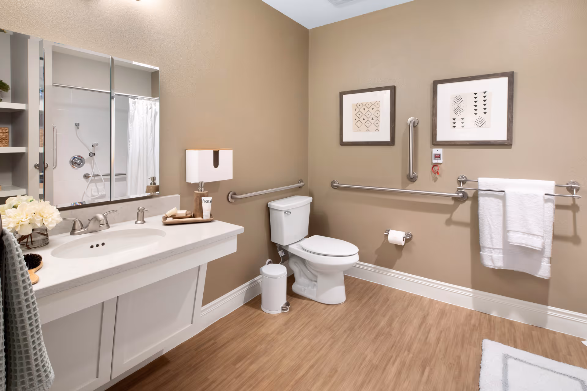 A clean and accessible bathroom with beige walls and wood-look flooring. The room features a white toilet with grab bars on the walls around it, a white vanity with a sink, soap dispenser, and decorative items, a large mirror above the sink, and a shower with a white curtain visible in the mirror. Two framed abstract artworks hang on the wall above the toilet, and a white towel is hanging on a towel rack.