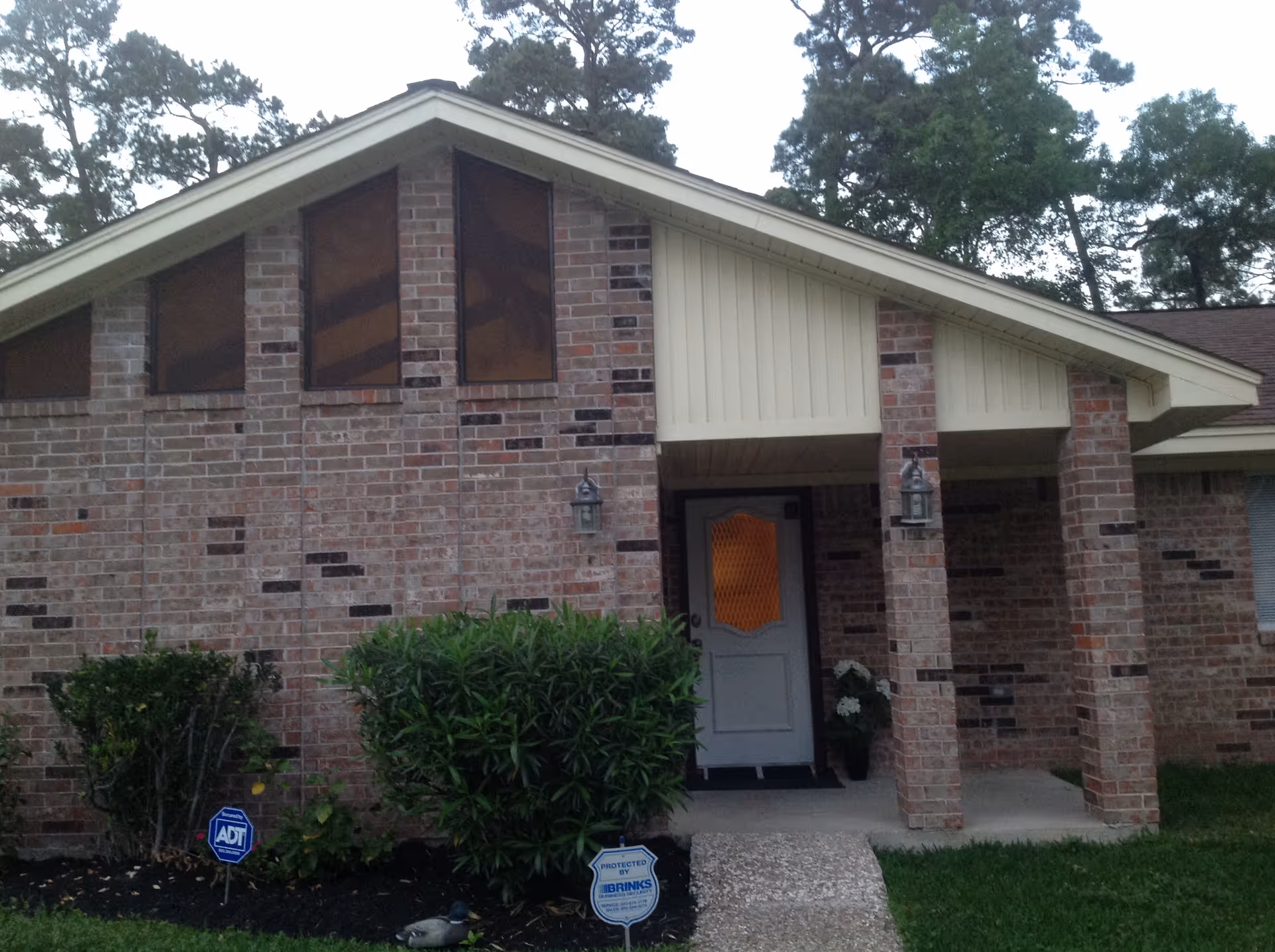 Exterior view of a brick building with a sloped roof and three triangular windows near the roof peak. There is a covered entrance with a white door featuring a decorative glass panel. Two lantern-style light fixtures are mounted on either side of the entrance. Shrubs and a small garden bed are in front of the building, along with security signs for ADT and Brinks.