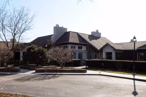 Exterior view of a single-story building with a sloped roof, surrounded by leafless trees and bushes, under a clear sky.