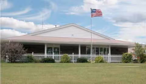 Single-story senior living facility with a white railing porch, shrubs, and American flag.