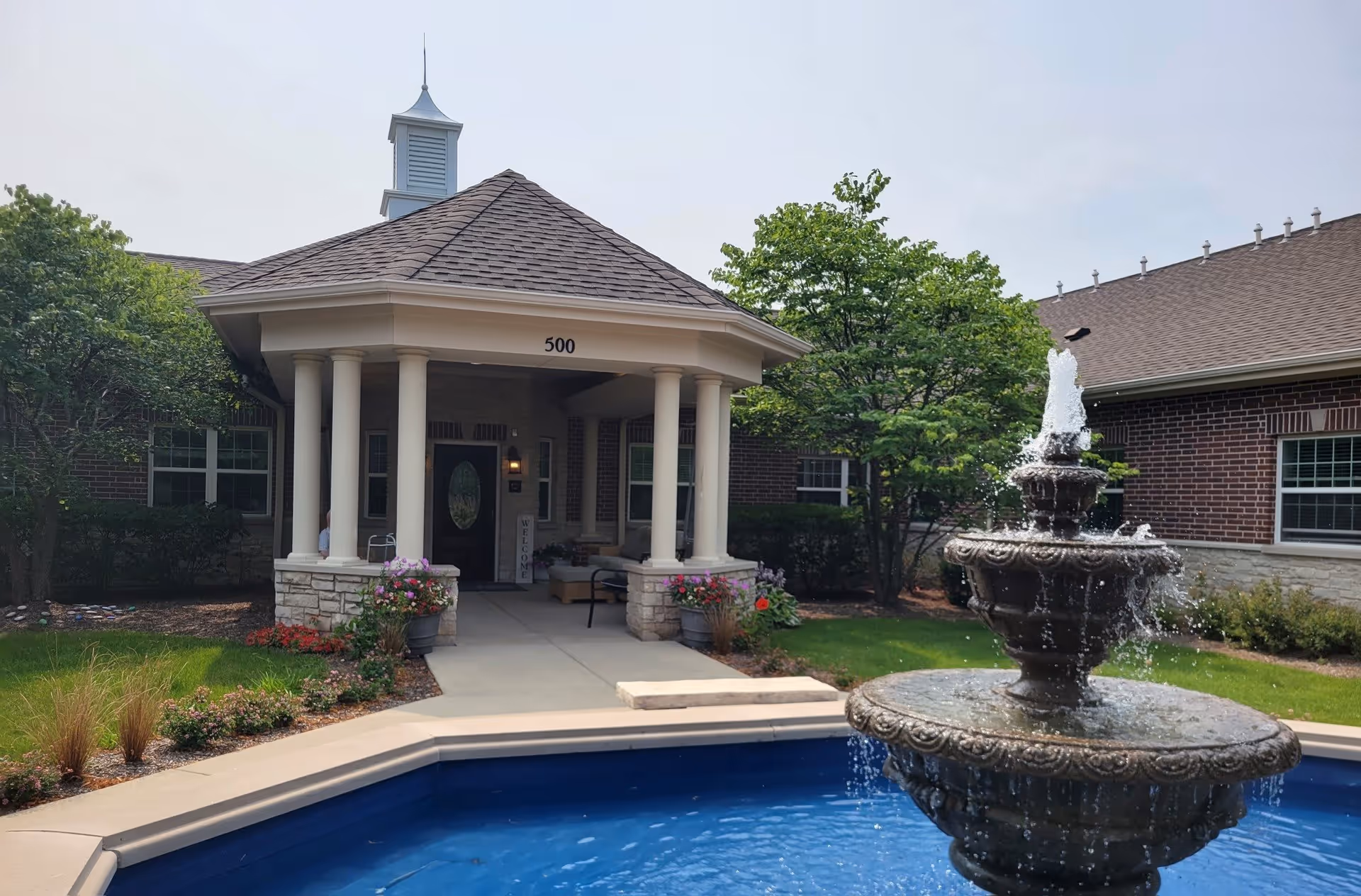 Entrance of a senior living facility with a covered porch supported by white columns, a decorative water fountain in the foreground, and well-maintained landscaping including trees and flowers.