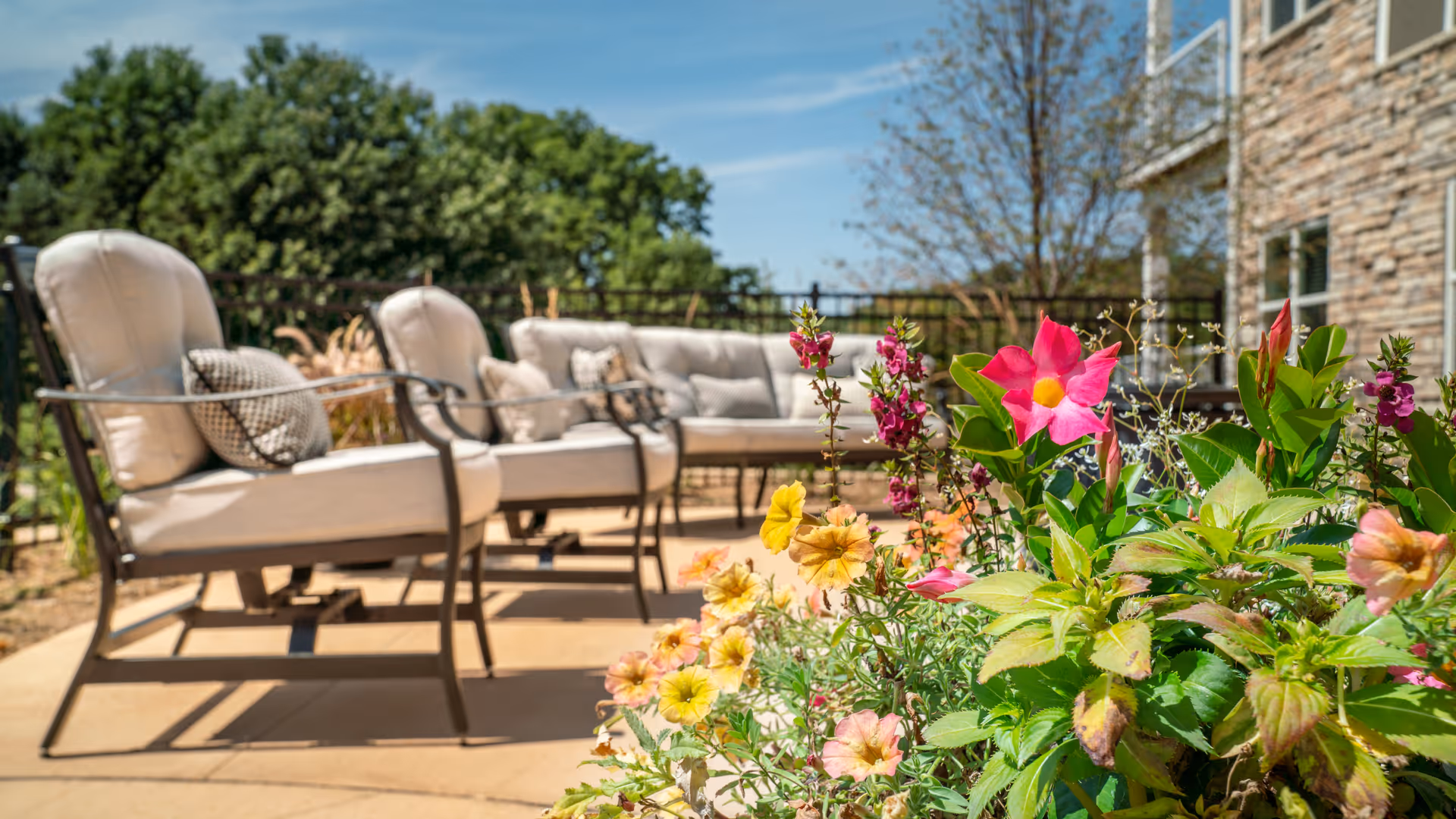 Outdoor patio area with cushioned metal chairs and a sofa arranged on a concrete surface, surrounded by colorful flowering plants and greenery under a clear blue sky.