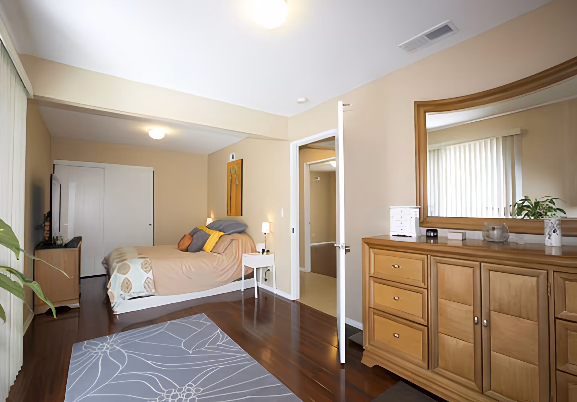 A bedroom with a bed against the wall, decorated with pillows and a blanket. There is a nightstand with a lamp beside the bed, a dresser with a large mirror, and a door leading to another room. The floor is wooden with a patterned rug, and there is a window with vertical blinds letting in natural light.