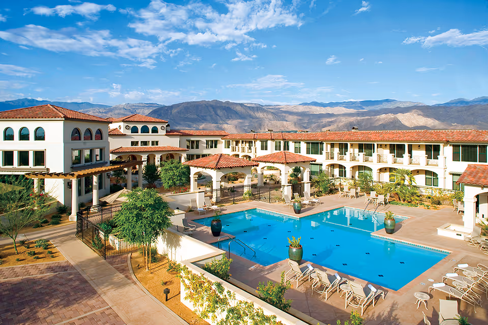 Outdoor view of Segovia senior living facility featuring a large swimming pool surrounded by lounge chairs and tables with umbrellas. The building has white walls and red tile roofs, with mountains visible in the background under a partly cloudy blue sky.