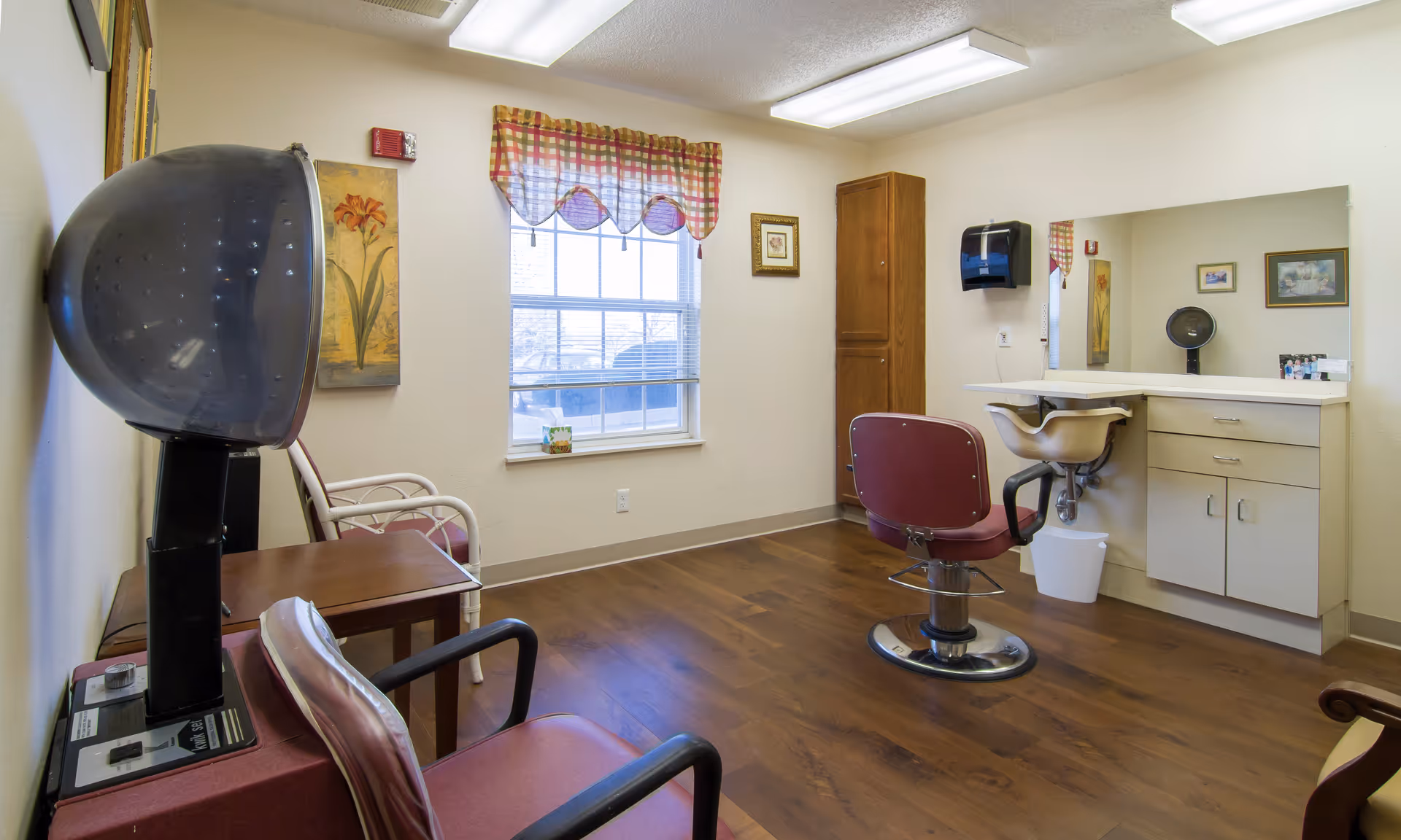 Small salon-style room with styling chairs, a hooded hair dryer, shampoo sink, mirror, window with valance, and cabinetry.