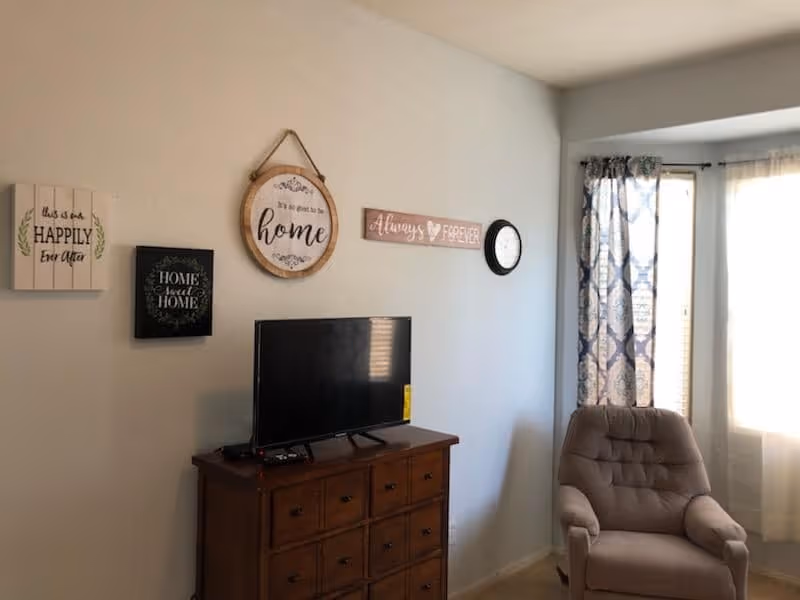 A cozy living room corner with a wooden dresser holding a flat-screen TV, a cushioned armchair next to a window with patterned curtains, and decorative wall signs with phrases like 'This is our happily ever after', 'Home sweet home', 'It's so good to be home', and 'Always & forever'.