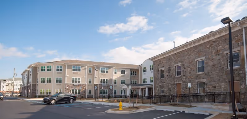 Exterior view of a senior living facility showing a multi-story building with beige and stone facade, multiple windows, a parking lot with a few cars, a yellow fire hydrant, and a clear blue sky with some clouds.