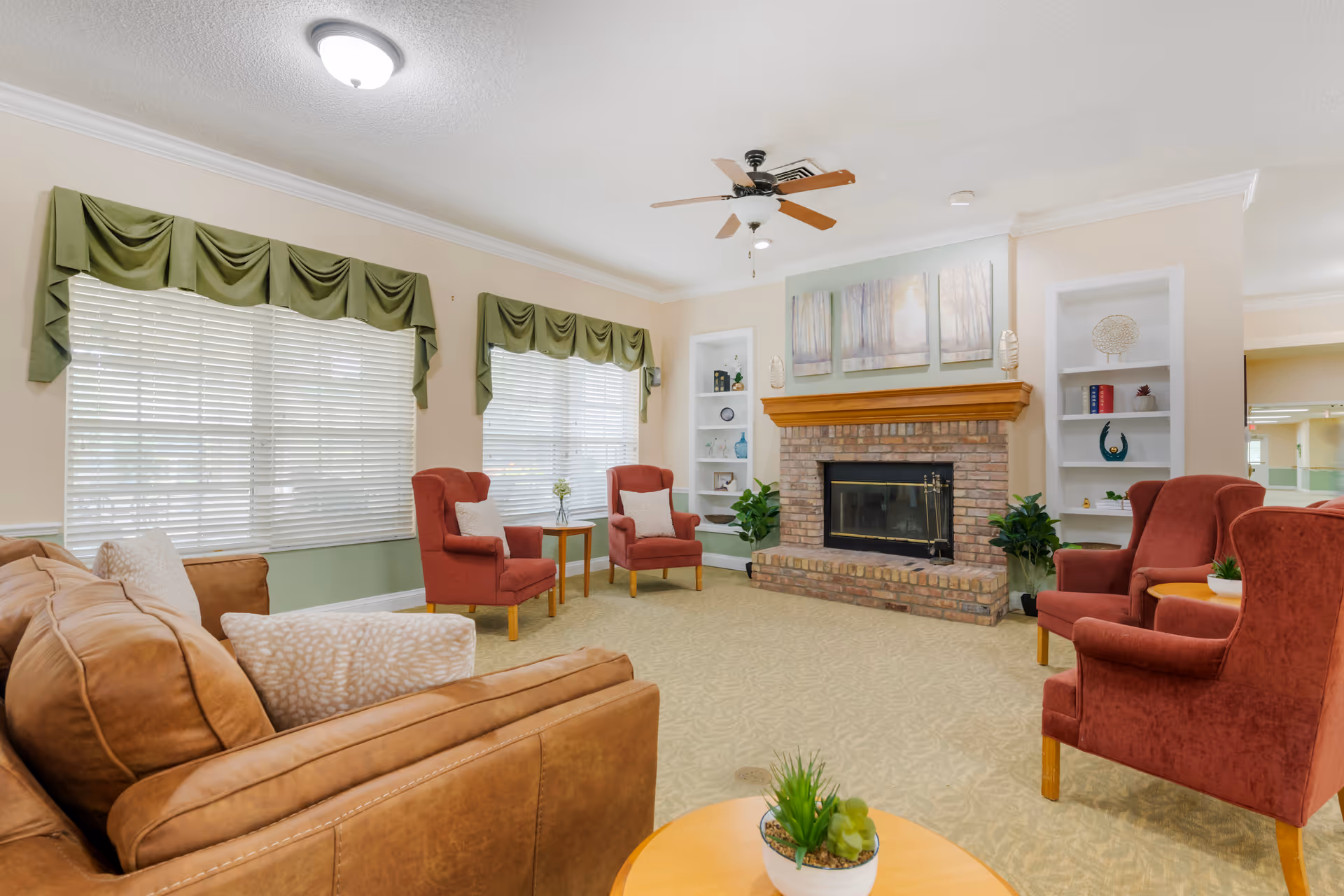 A cozy living room in a senior living facility featuring a brown leather sofa with patterned pillows, four red upholstered armchairs, a brick fireplace with a wooden mantle, two built-in white shelves with decorative items, two large windows with green valances, a ceiling fan, and a round wooden coffee table with a small potted plant.