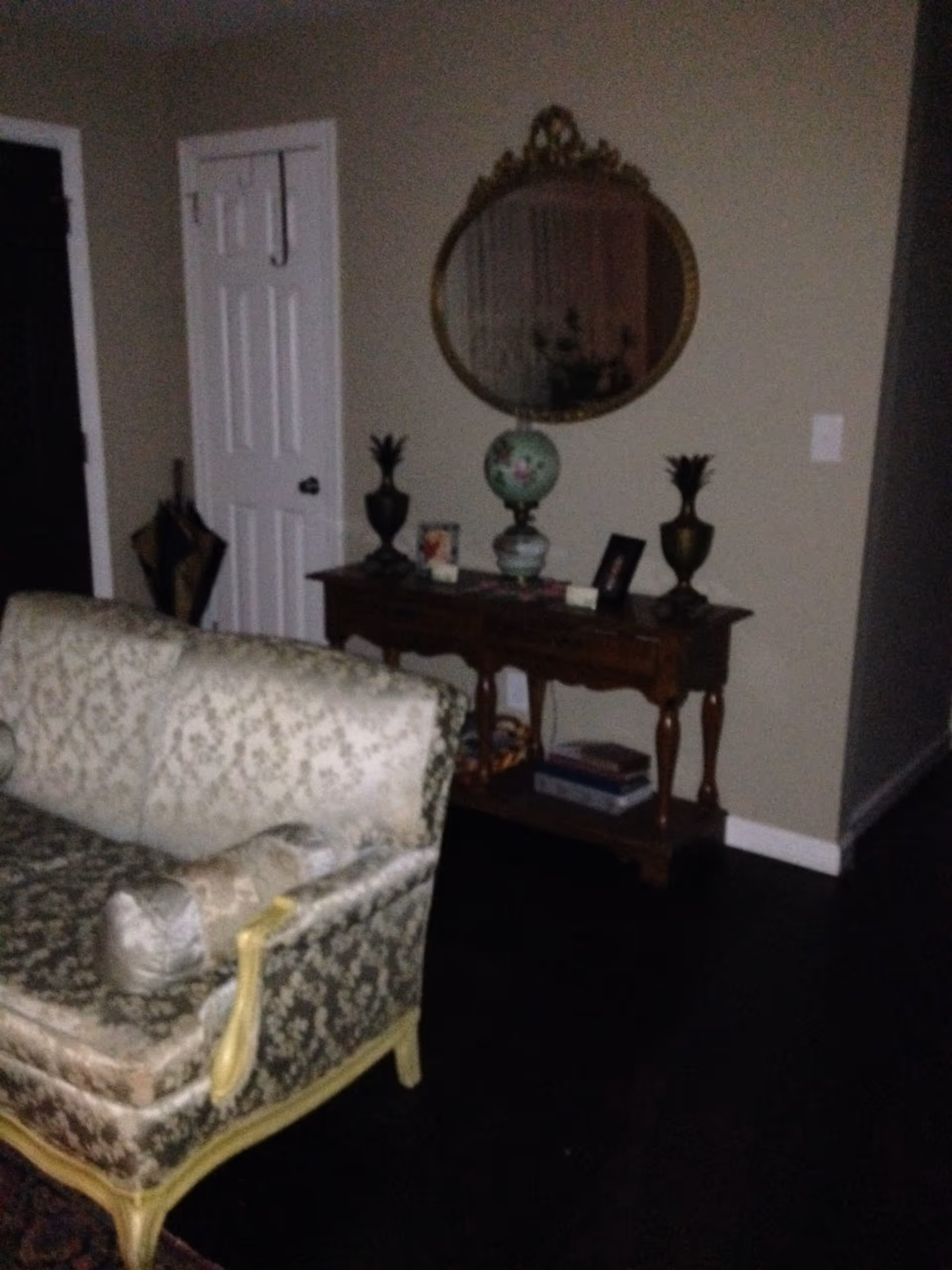 A dimly lit living room area featuring a vintage-style sofa with patterned upholstery and a wooden console table against the wall. The table holds decorative items including a floral lamp, two pineapple-shaped ornaments, and framed photos. Above the table is an ornate oval mirror. A closed white door and a dark umbrella stand are visible in the background.