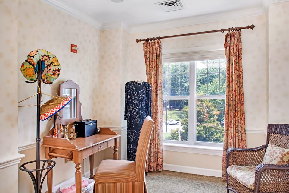 A cozy interior corner of a room featuring a wooden vanity table with a mirror, a striped chair, a coat rack with hats, a mannequin dressed in a dark floral dress, and a large window with patterned curtains overlooking greenery outside. There is also a wicker armchair with a patterned cushion.
