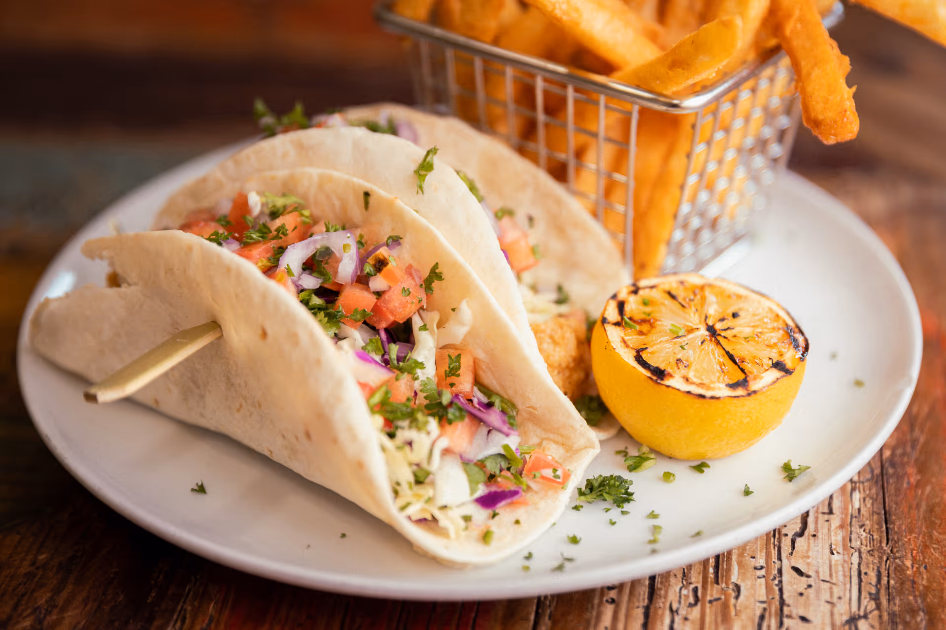 Close-up of two soft tacos filled with chopped tomatoes, onions, cabbage, and herbs on a white plate, served with a grilled lemon half and a small metal basket of French fries on a wooden table.
