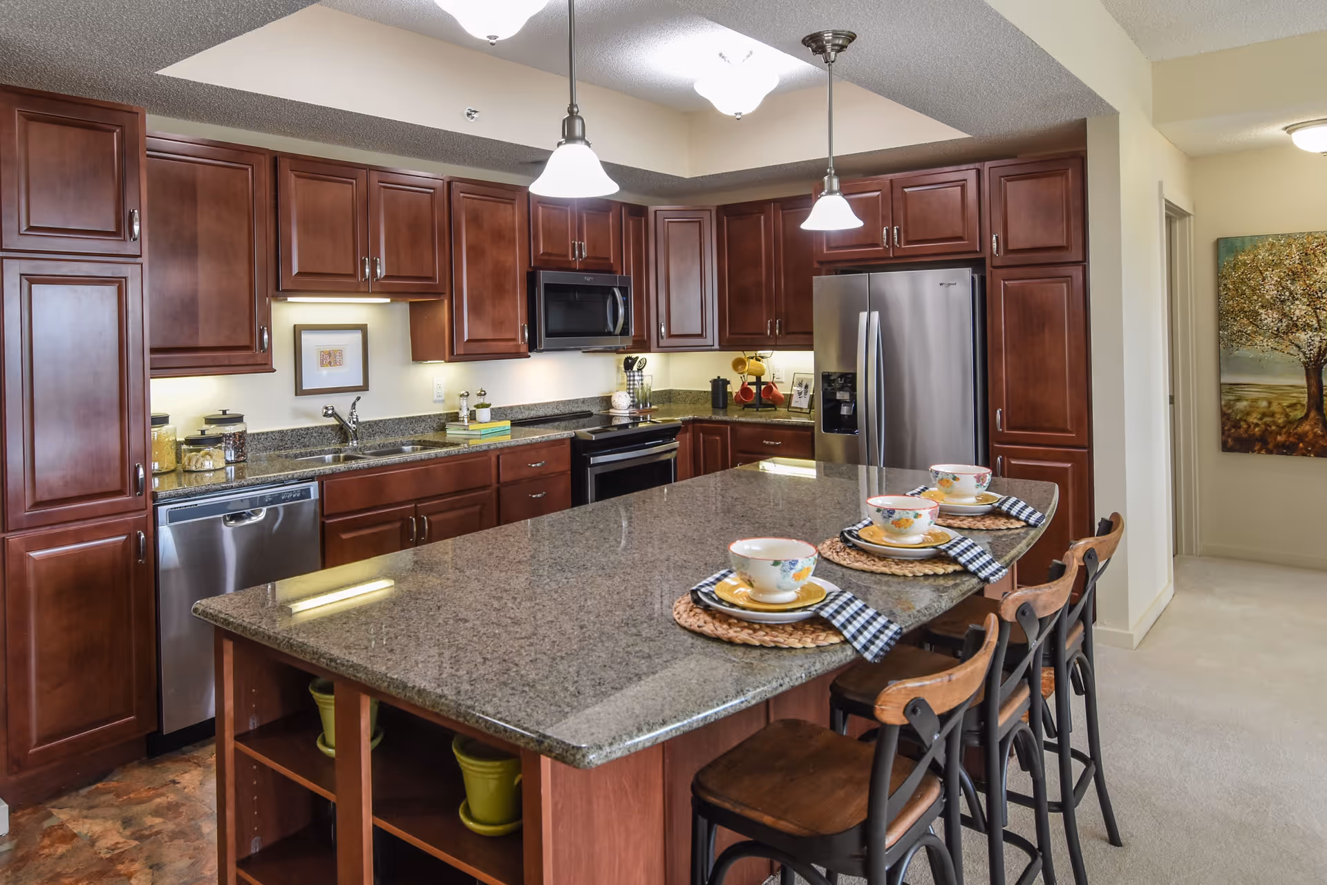 A modern kitchen with dark wood cabinets, stainless steel appliances including a refrigerator, dishwasher, and microwave. There is a large granite island in the center with three place settings, each with a bowl, plate, and napkin. Three wooden bar stools are positioned at the island. The kitchen is well-lit with pendant lights hanging above the island and recessed lighting.