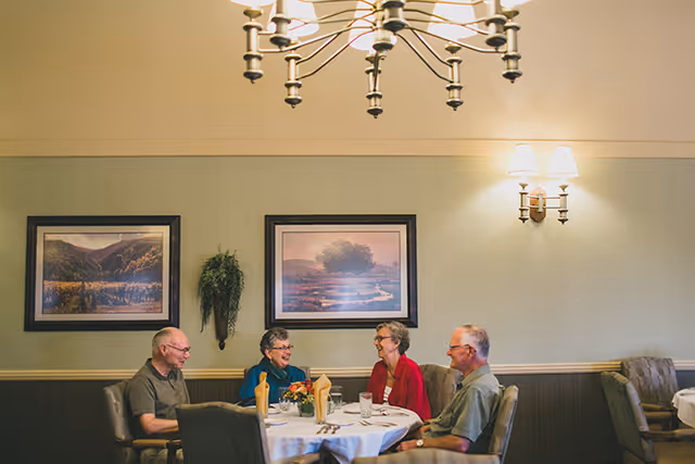 Four older adults seated around a round dining table in a well-lit communal dining room, chatting and smiling.