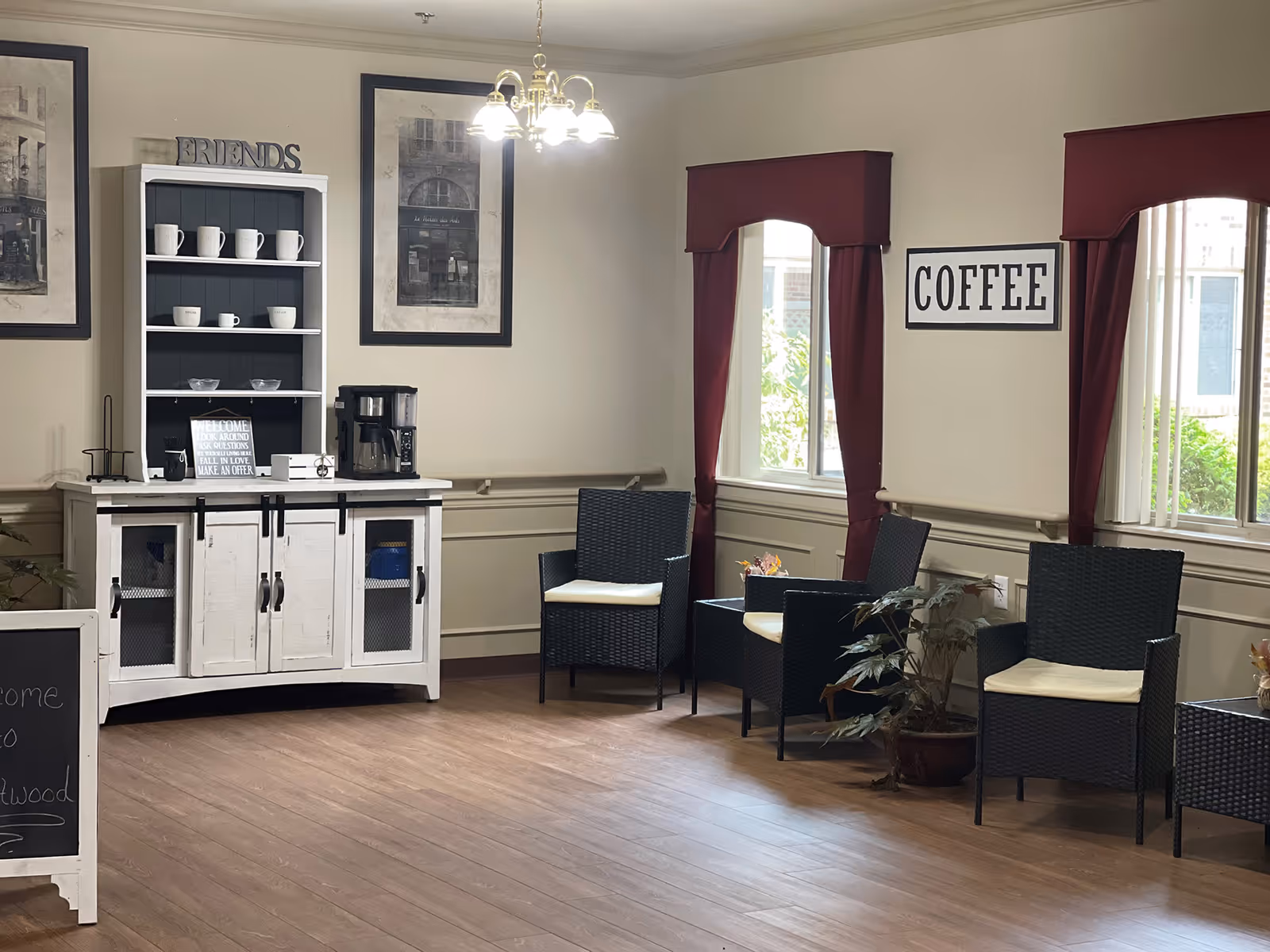 A cozy seating area in a senior living facility with three black wicker chairs with white cushions arranged around a small table. There is a white cabinet with shelves holding white mugs and a coffee maker. The walls are decorated with framed pictures and a sign that says 'COFFEE'. Two windows with red curtains let in natural light.