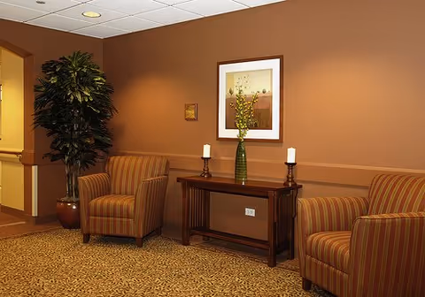 A cozy seating area with two striped armchairs on either side of a wooden console table. The table holds a green vase with yellow flowers and two white candles on candle holders. A framed artwork hangs on the brown wall above the table, and a large potted plant is placed in the corner near an open doorway.