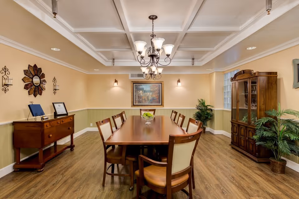 A well-lit dining room with a long wooden table surrounded by eight chairs. The room features a coffered ceiling with two chandeliers, beige walls with green wainscoting, wooden flooring, a sideboard with decorative items, a glass-fronted cabinet, potted plants, and a framed painting on the far wall.