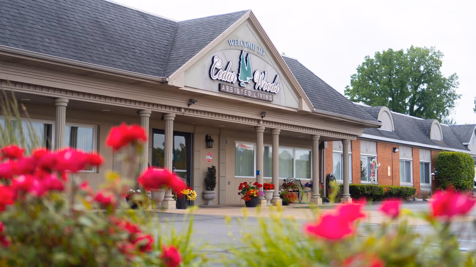 Front exterior view of Cedar Woods Assisted Living & Memory Care facility with a covered entrance supported by columns, flower pots with colorful flowers, and a sign above the entrance that reads 'Welcome to Cedar Woods Assisted Living'.