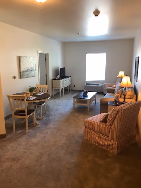 A cozy living room area with beige carpet, a striped armchair, a sofa with cushions, a coffee table, and a TV on a white stand. There is a small round dining table with two chairs on the left side, a window letting in natural light, and two table lamps providing warm lighting.