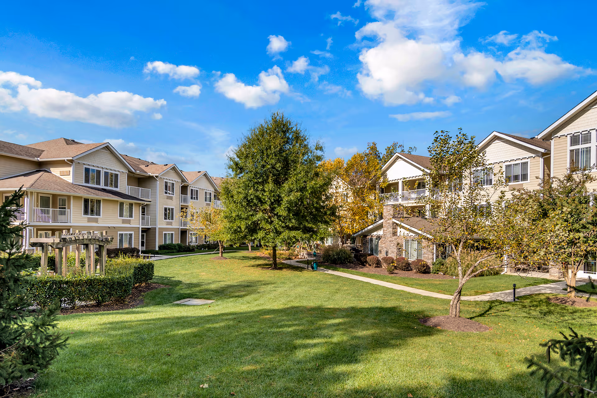A bright outdoor view of Chesterfield Heights Gracious Retirement Living featuring well-maintained green lawns, trees, and shrubs. The image shows two beige multi-story residential buildings with balconies under a partly cloudy blue sky.