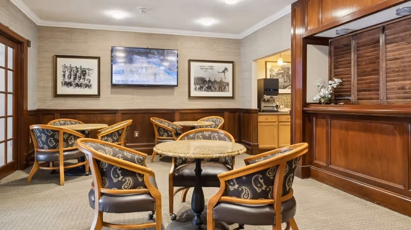 A cozy seating area with round tables and cushioned chairs featuring a paisley pattern. The room has wood paneling on the walls, two framed black and white pictures, and a flat-screen TV mounted on the wall. There is a small counter area with wooden shutters and a coffee machine in an adjacent space.