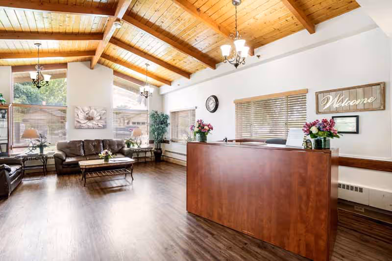 A spacious reception area with a wooden ceiling and beams, featuring a wooden reception desk adorned with flower arrangements. Behind the desk is a wall with a clock and a 'Welcome' sign. The room has large windows with blinds, brown leather sofas, a coffee table, side tables with lamps, and potted plants, creating a warm and inviting atmosphere.